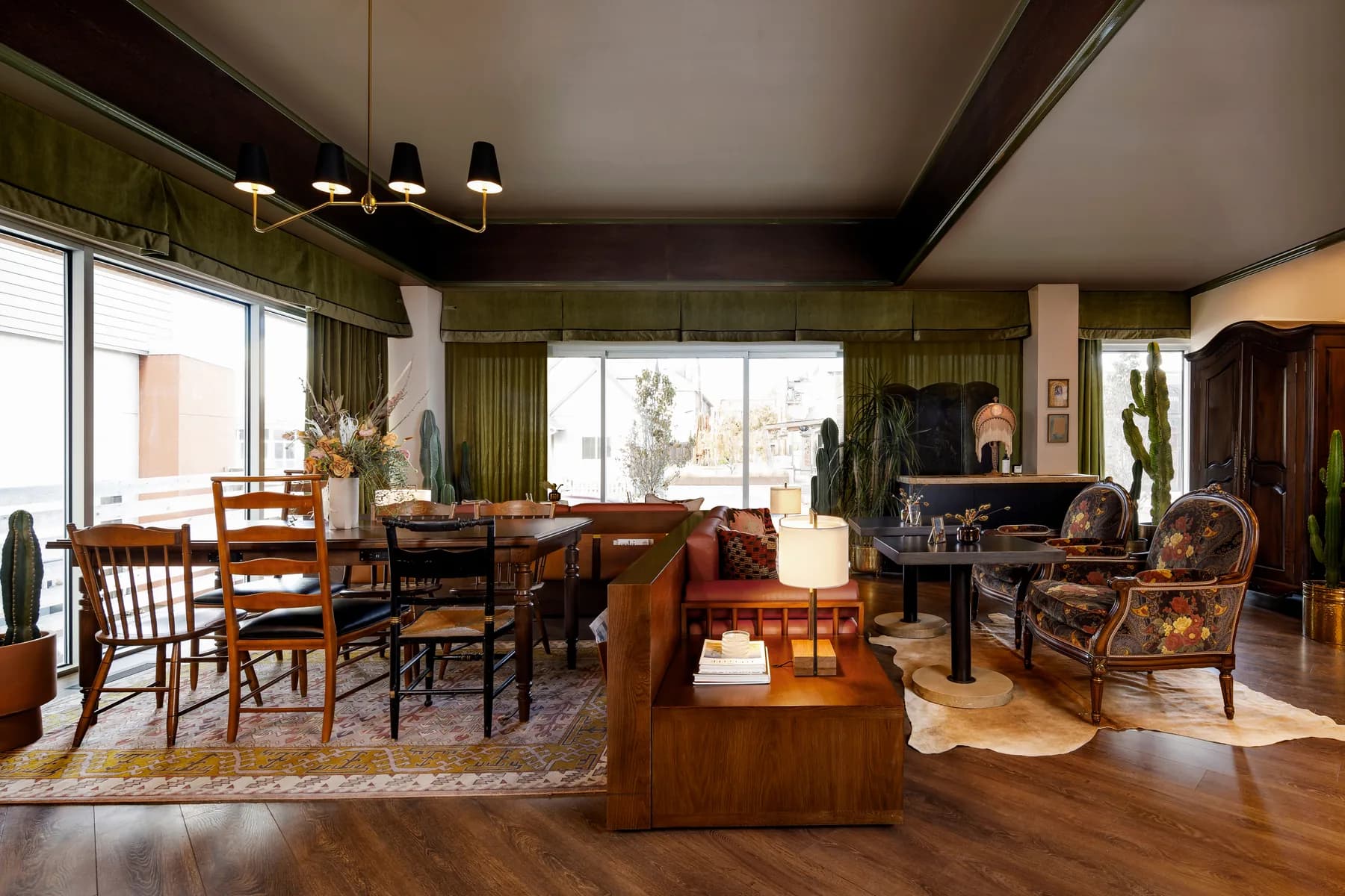 A sunlit living room at Wildflower with green drapes, communal tables, a leather banquette and floral armchairs.